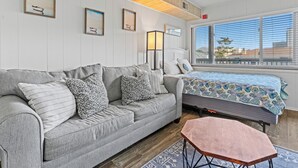 Ein Zimmer mit grauem Sofa, einem kleinen Holztisch, einem Bett mit gemustertem Überwurf und einem Fenster mit Jalousien.