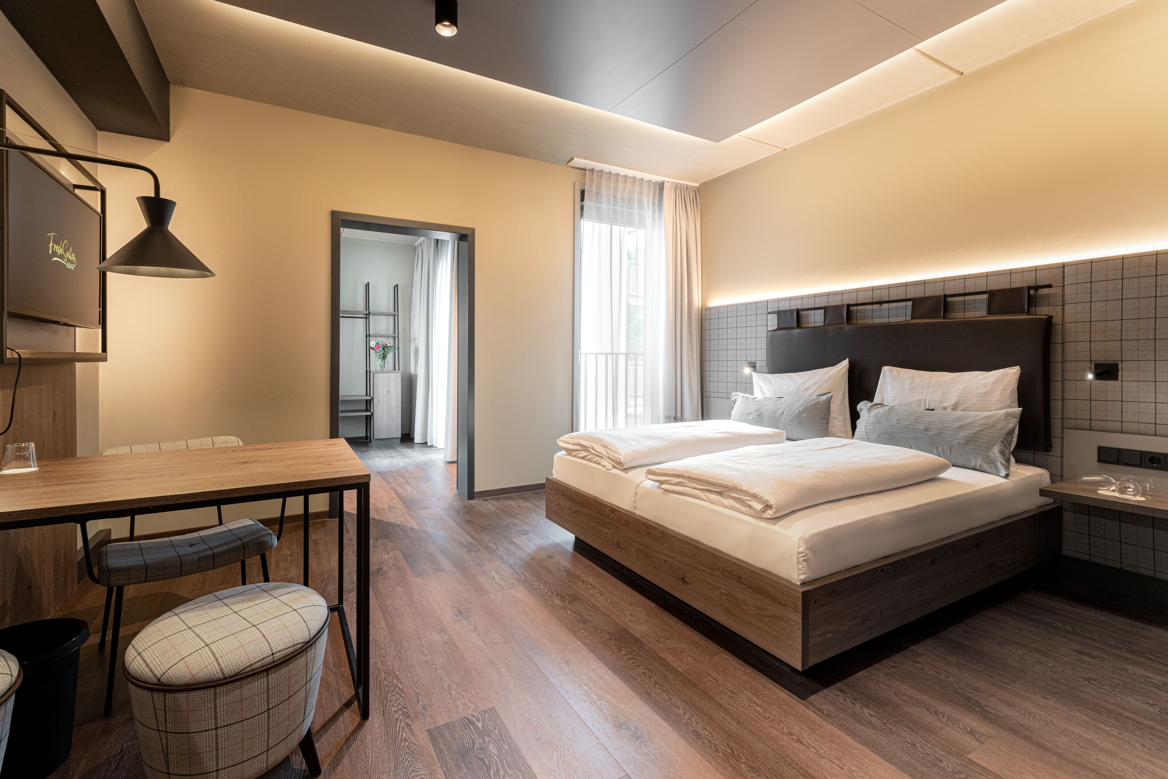 A modern hotel room with a wooden floor, a large bed with white linens, a desk with a chair, and a small table with a glass.