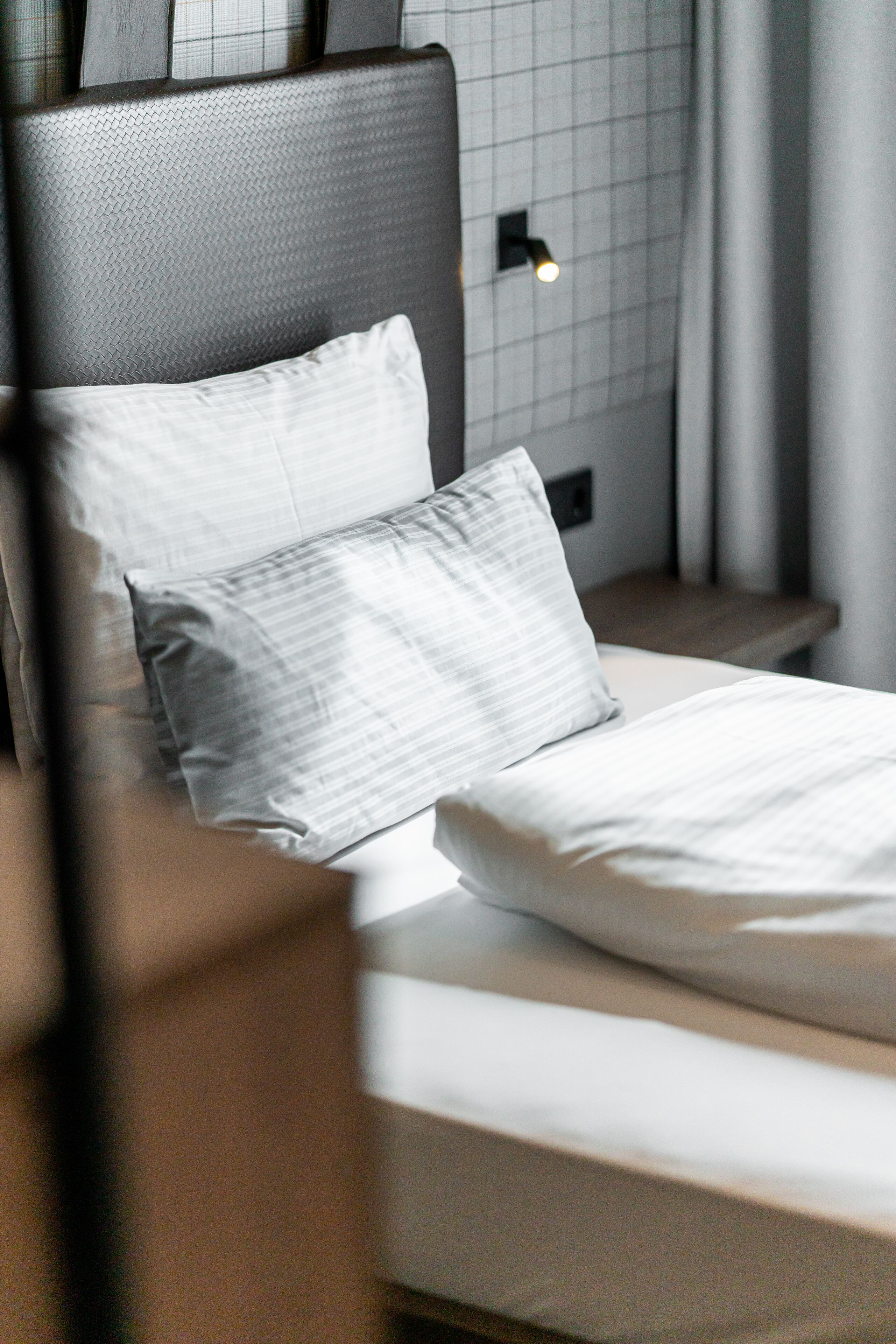 Een net opgemaakt bed met witte lakens en twee kussens, een hoofdbord met een structuurpatroon, en een nachtkastje met een kleine lamp.