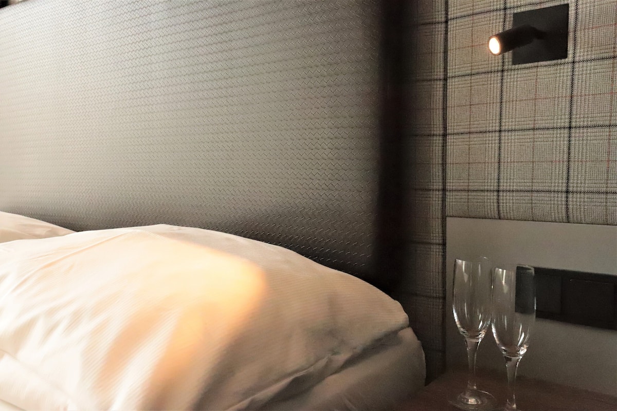 A hotel room with a textured headboard, a bedside table with two empty wine glasses, and a wall-mounted light fixture.