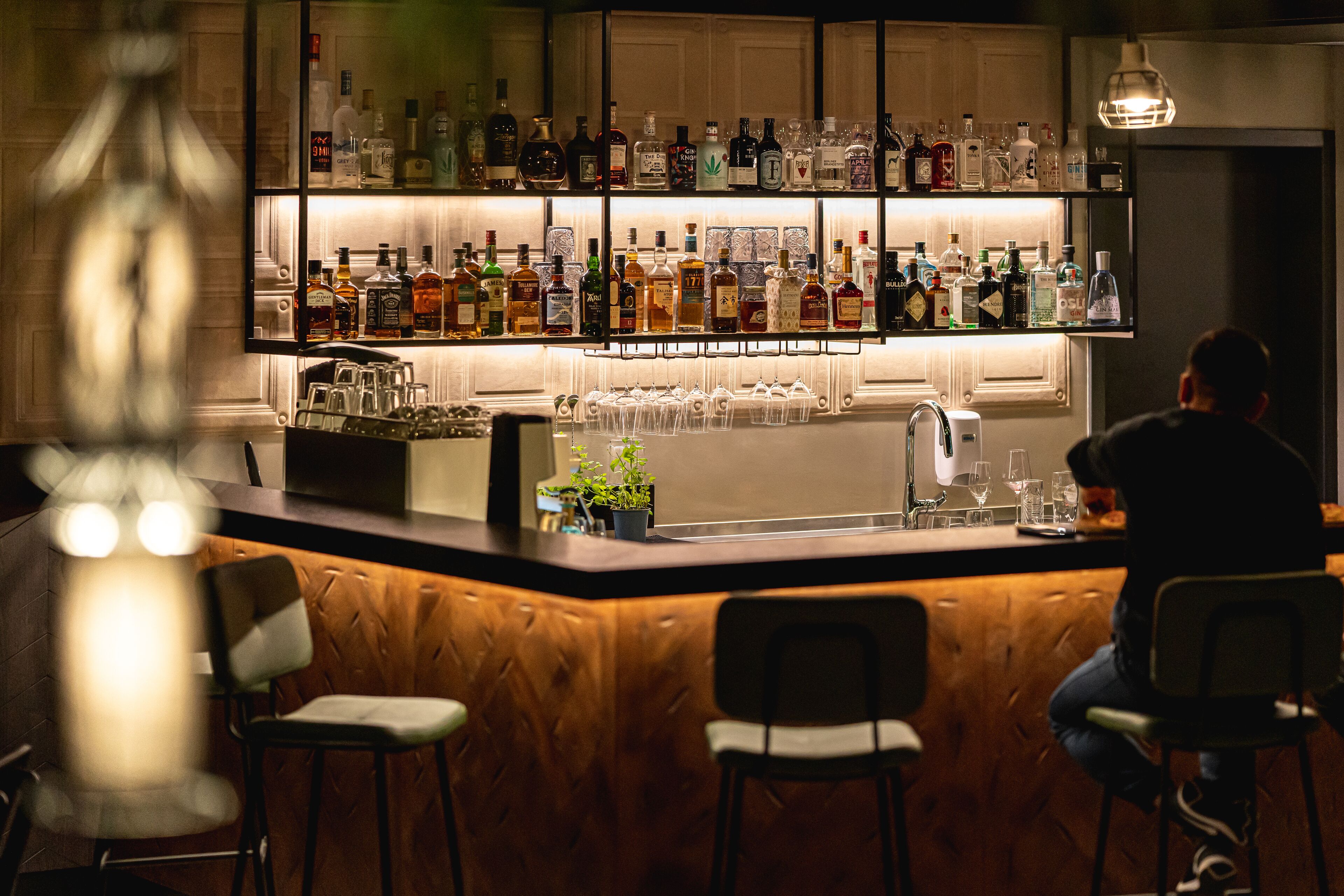 Zona de bar con diferentes botellas de licor y vasos. Hay dos taburetes y una planta en maceta sobre la barra.