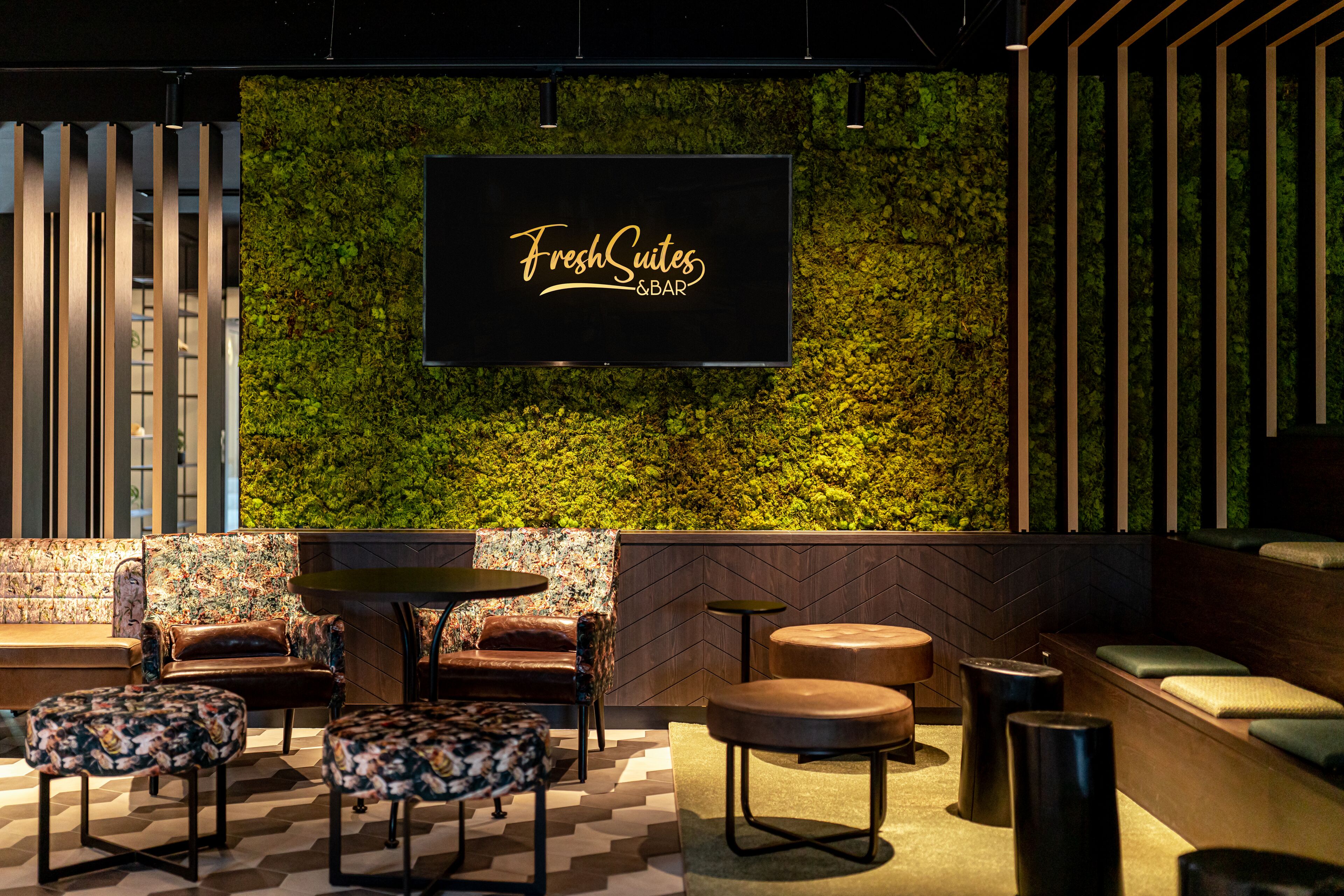 Uma área de lounge moderna com uma parede verde, uma televisão de tela plana exibindo "FreshSuites & BAR" e assentos confortáveis, incluindo sofás e pufes.