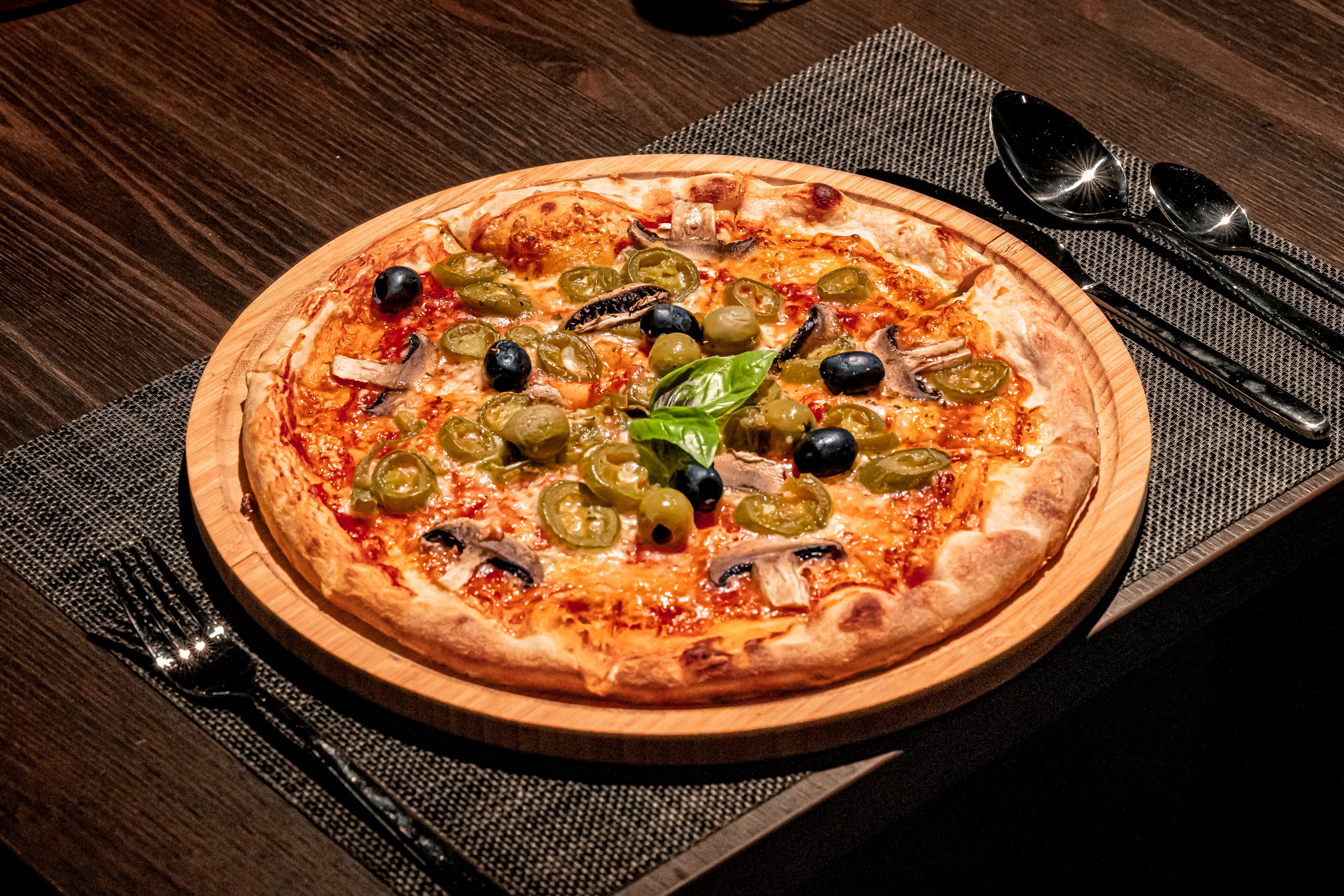A pizza on a wooden plate with olives, green peppers, and basil on a table.