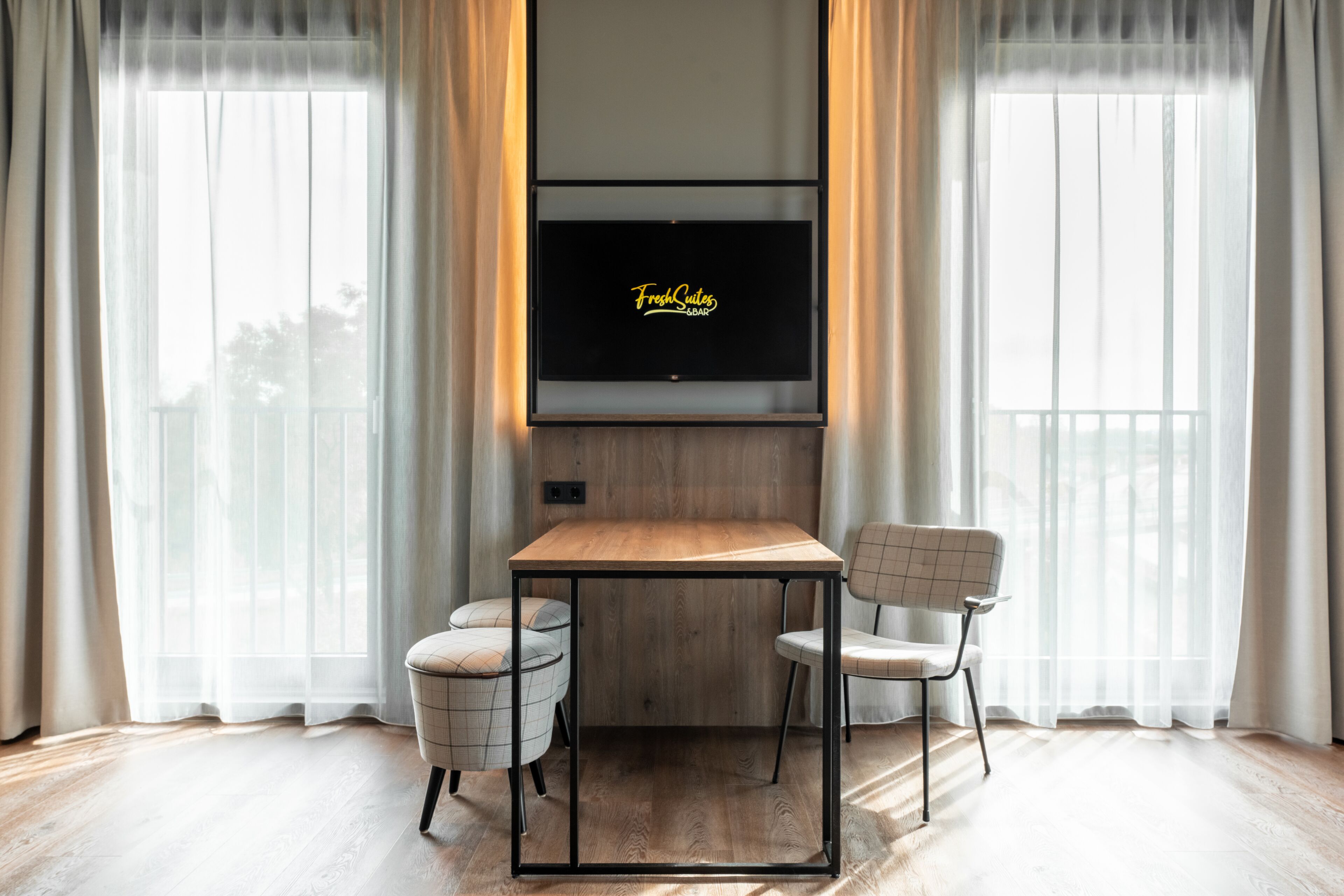 A modern room with a wooden table and two chairs, a flat-screen TV mounted above the table, and sheer curtains on the windows.