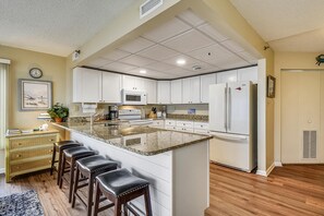 Une cuisine avec des meubles blancs, un îlot central doté de tabourets, un réfrigérateur et une horloge au mur.