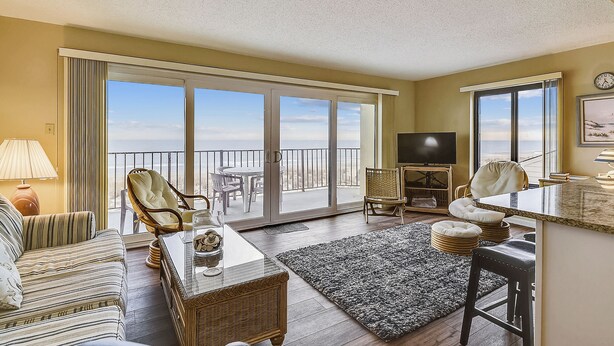 Una sala de estar con sofá, sillas, televisor, balcón con vistas y comedor.