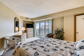 Une chambre à coucher comprenant un lit, une commode, une chaise, un téléviseur et une fenêtre donnant sur des immeubles.