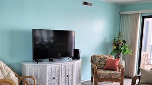 Uma sala de estar com uma televisão de tela plana sobre um armário branco, cadeiras de vime, uma mesa de centro de vidro e um vaso de planta.