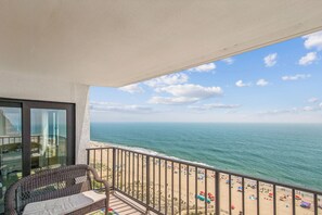 Un balcon avec vue sur l’océan et la plage, des meubles en osier et une balustrade.