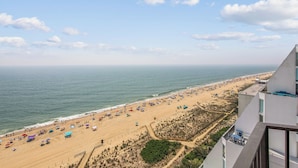 A beach view with a clear sky, sandy beach, and a building with a balcony overlooking the beach. - XPLORIE ACTIVITIES & LINENS INCLUDED*! Plaza 3BR/2BA END UNIT with oceanfront & bayside balconies! (Ocean City)