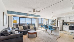 A modern living room with a sofa, armchair, and round coffee table. There is a dining area with blue chairs and a kitchen with white cabinets. - XPLORIE ACTIVITIES & LINENS INCLUDED*!  OCEAN & BAY VIEWS!! 2BR Capri Condo on 110th Street. (Ocean City)