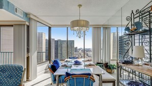 A dining area with a table set for two, a chandelier, and a view of the cityscape through large windows. - XPLORIE ACTIVITIES & LINENS INCLUDED*!  OCEAN & BAY VIEWS!! 2BR Capri Condo on 110th Street. (Ocean City)