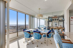 Une pièce spacieuse avec une grande table à manger, des chaises recouvertes d’un tissu bleu et une vue sur le paysage urbain grâce à de grandes fenêtres.
