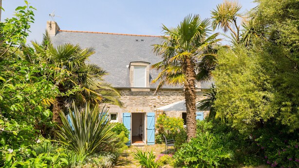 Une maison en pierre avec des volets bleus, entourée d’une végétation luxuriante et d’un jardin.