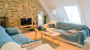 Un salon chaleureux avec un mur en pierre, un téléviseur, un canapé, une table basse et une fenêtre laissant entrer la lumière naturelle.