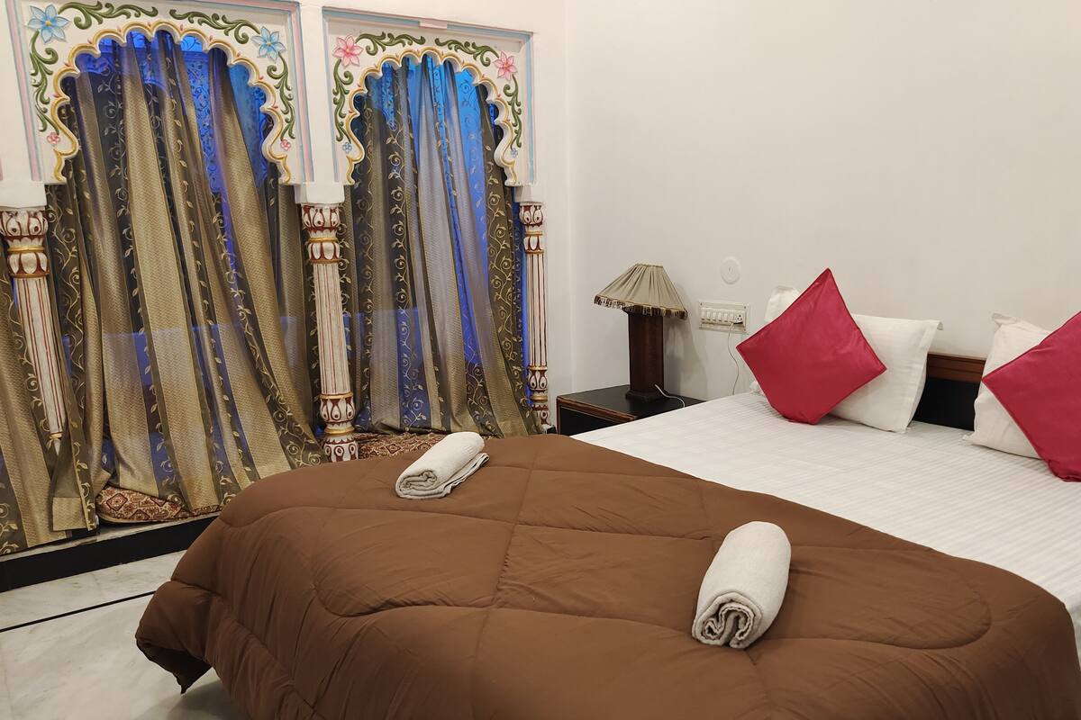 A hotel room with a large bed, red pillows, a nightstand with a lamp, and ornate curtains.