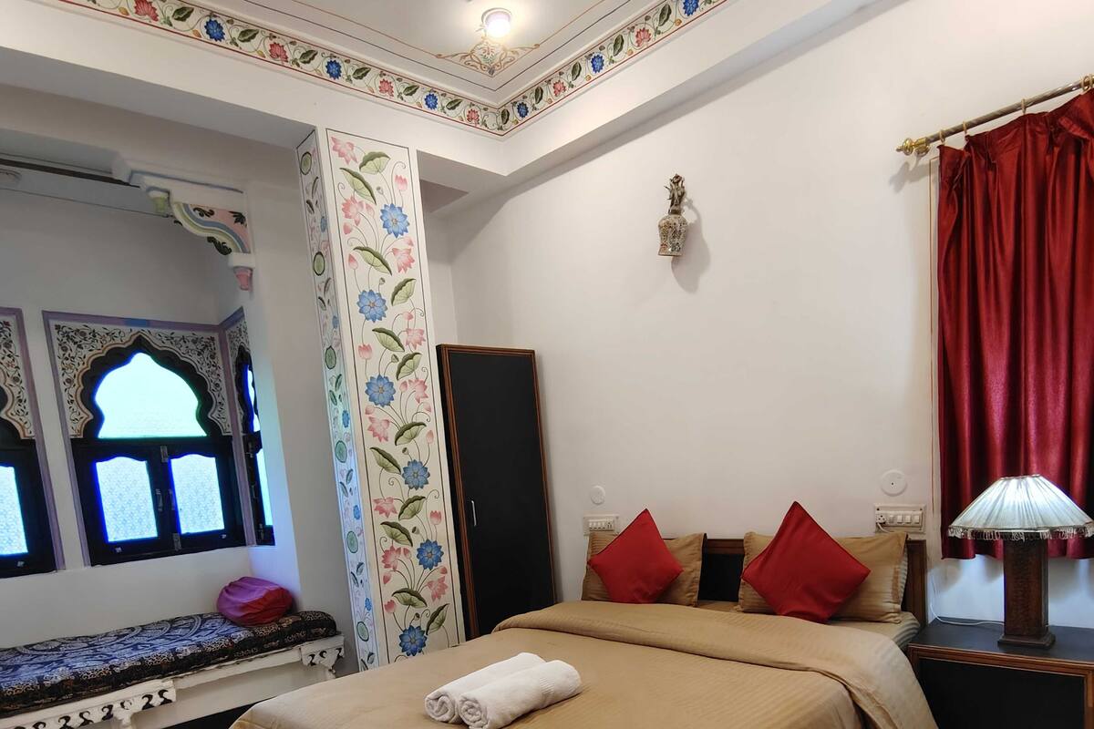 A bedroom with a bed, pillows, a nightstand, a lamp, a window with floral design, and a ceiling fan.