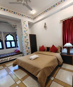 A bedroom with a bed, pillows, a nightstand, a lamp, a window with floral design, and a ceiling fan. - The Rustic Monk (Udaipur)