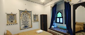 A room with a bed, a wardrobe, a window with blue curtains, and decorative wall art. - The Rustic Monk (Udaipur)