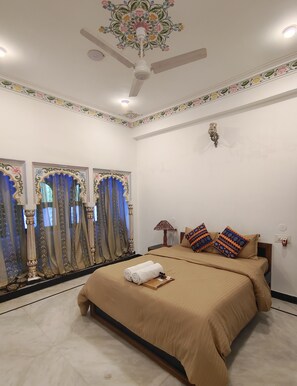 A bedroom with a large bed, two pillows, a bedside table with a lamp, a ceiling fan, and decorative curtains. - The Rustic Monk (Udaipur)