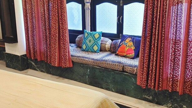 A room with a bed, a window with patterned curtains, a bench with cushions, and a checkered floor. - The Rustic Monk (Udaipur)