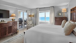 A hotel room with a large bed, a TV, a desk with a lamp, a chair, and a view of the sea through the window.