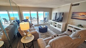 A modern living room with a large TV, comfortable seating, and a view of the ocean through large windows. - Sandy Key 438: 3 BR/2 BA Beachfront Condo, Perdido Key, FL. On-Site Rental Office, Sleeps 8 (Pensacola)