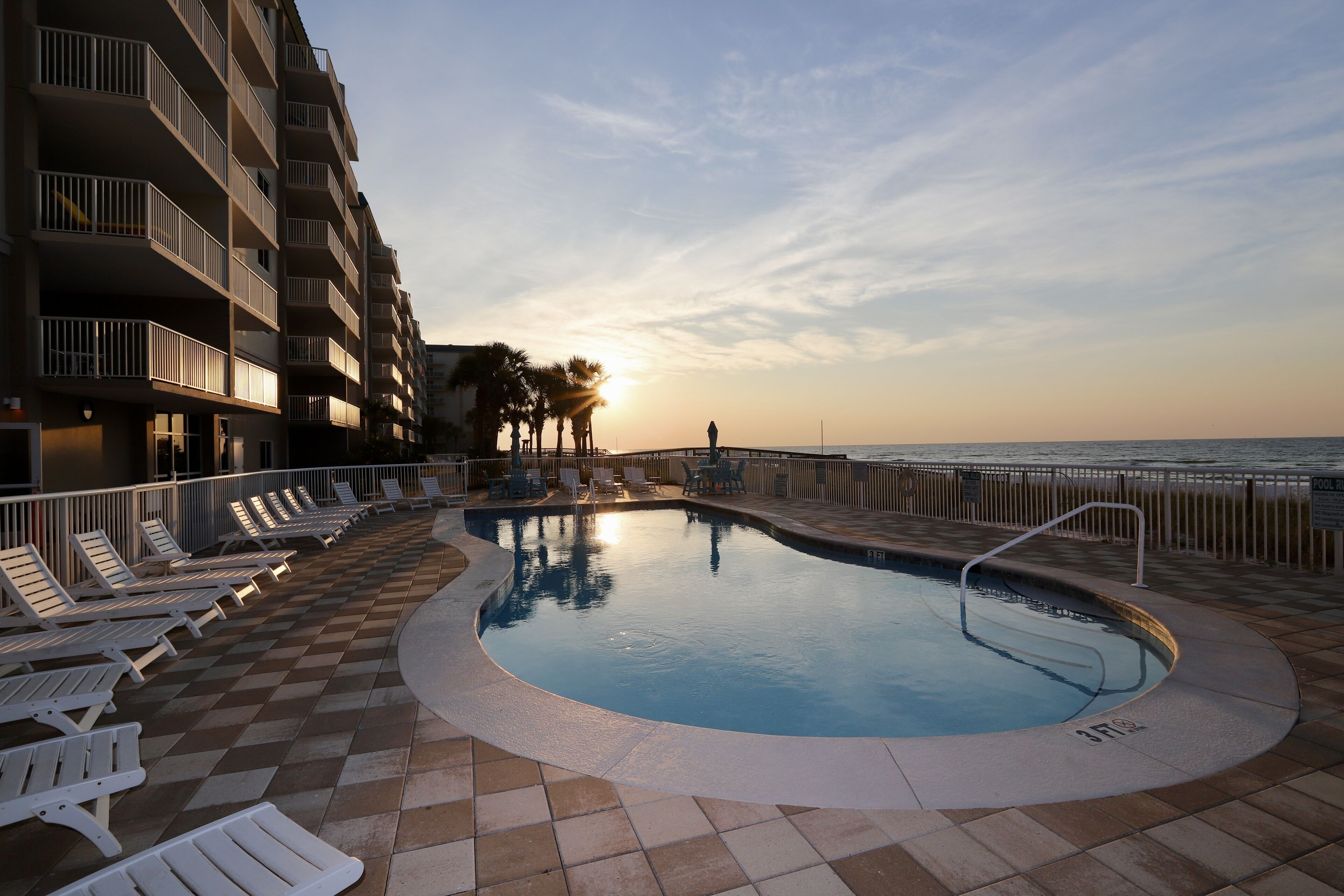 A pool area with lounge chairs and a view of the ocean at sunset.