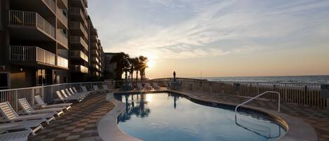A pool area with lounge chairs and a view of the ocean at sunset.