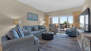 Ein geräumiges Wohnzimmer mit einer Sofagarnitur, Hocker und einem Flachbildfernseher. Durch die Glastüren ist ein Balkon mit Gartenmöbeln zu sehen.