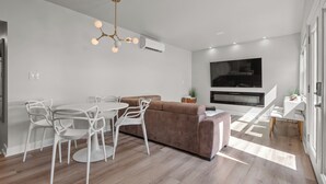 Una sala de estar moderna con un sofá marrón, sillas blancas, una mesa de comedor y un televisor montado en la pared.