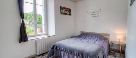 A bedroom with a bed, a window with curtains, a radiator, a wall decoration, and a bedside lamp.