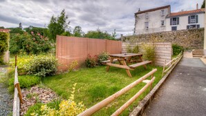 A garden with a wooden table and benches, a stone wall, and a building in the background. - Gite Le Mistral 2/4 persons 6kms from the puy du fou park (Mallièvre)