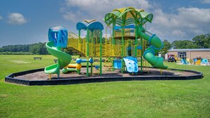 A playground with slides and climbing structures, surrounded by a grassy field and a building in the background. - Waterview Cabin on Currituck Sound - Great Campground Amenities (WV90) (Coinjock)