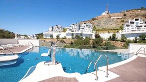 A swimming pool with a wooden deck, lounge chairs, and a view of hillside buildings. - La Quinta 3 bedroom apartment with Private Hot Tub (Benahavís)