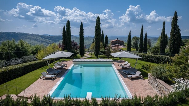 Une piscine rectangulaire bordée de chaises longues et d’ombrelles, entourée de pelouses bien entretenues et de grands cyprès, avec une chaîne de montagnes en toile de fond.