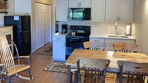 Una cocina moderna con armarios blancos, un refrigerador negro, microondas y cafetera. Hay una mesa de comedor de madera con sillas y un sillón mecedora.