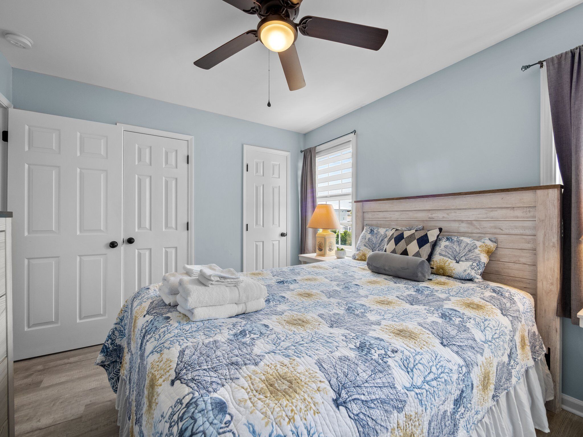 A bedroom with a bed, a ceiling fan, a window with curtains, and a nightstand with a lamp.