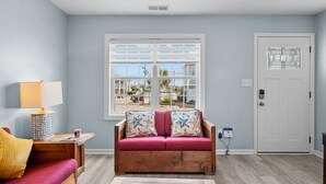 A living room with a wooden sofa, a red armchair, a lamp, and a window with a view of houses. - Oceanside Condo in Atlantic Beach, NC-Anchors Away Unit 3 (Atlantic Beach)