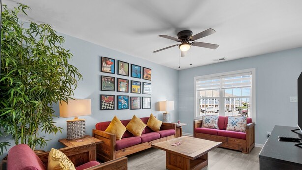 A modern living room with a sofa set, a coffee table, a side table with a lamp, a large potted plant, and a ceiling fan. - Oceanside Condo in Atlantic Beach, NC-Anchors Away Unit 3 (Atlantic Beach)
