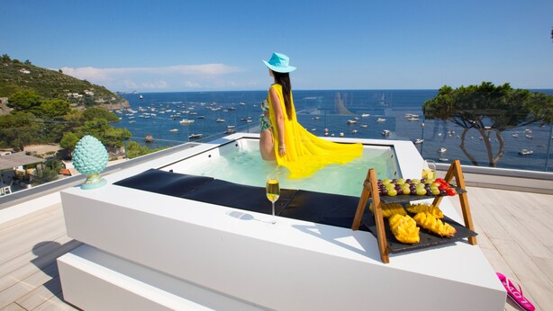 Une femme vêtue d’une robe jaune et d’un chapeau bleu est assise dans un bain à remous, d’où elle peut admirer les bateaux et la mer.