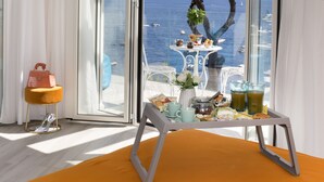 Une chambre avec une grande fenêtre donnant sur la mer, une table avec des aliments pour le petit-déjeuner et un petit tabouret orange.
