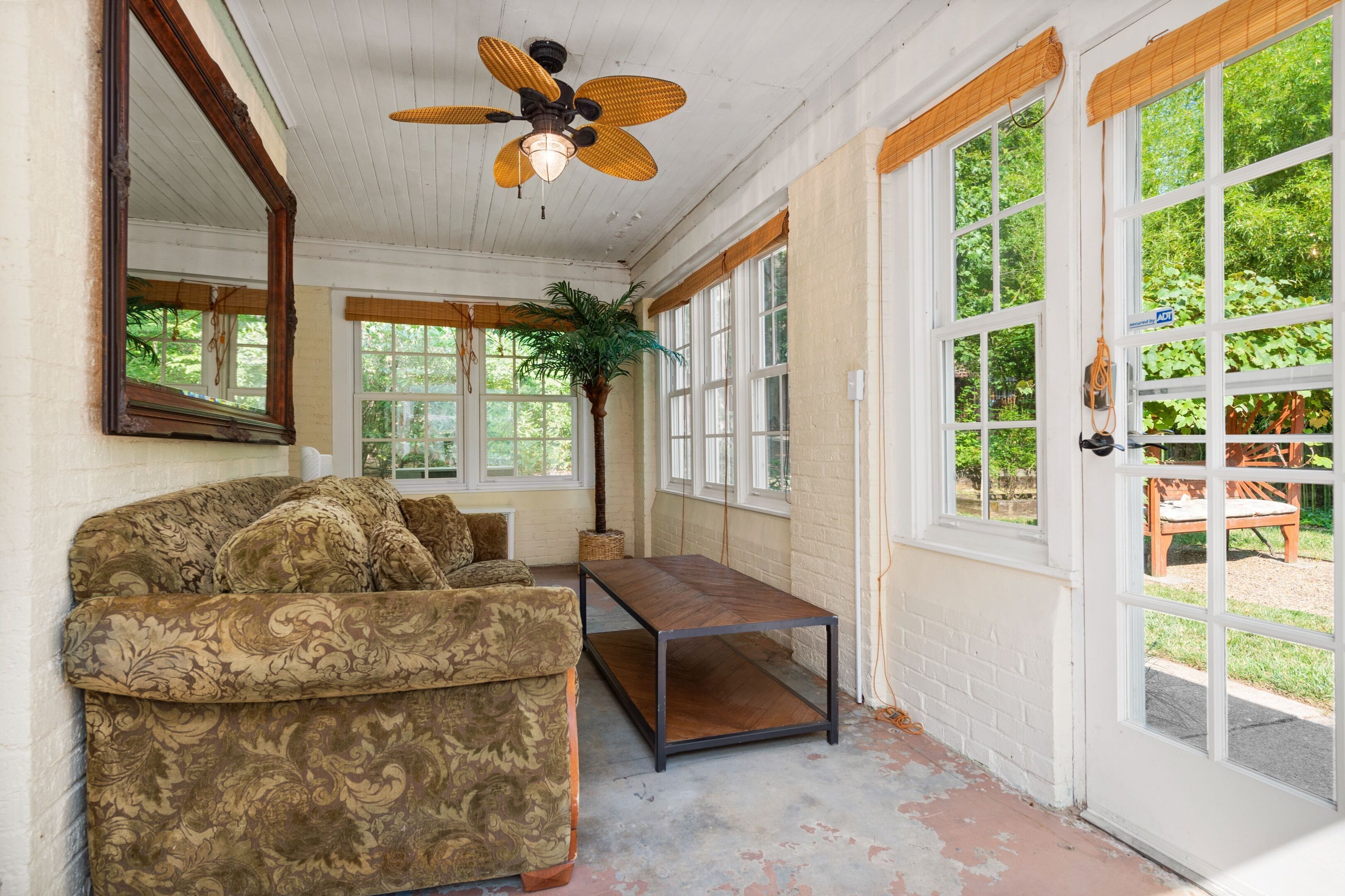 Une pièce avec un canapé à motifs, une table basse en bois, un ventilateur de plafond et de grandes fenêtres donnant sur un paysage verdoyant.
