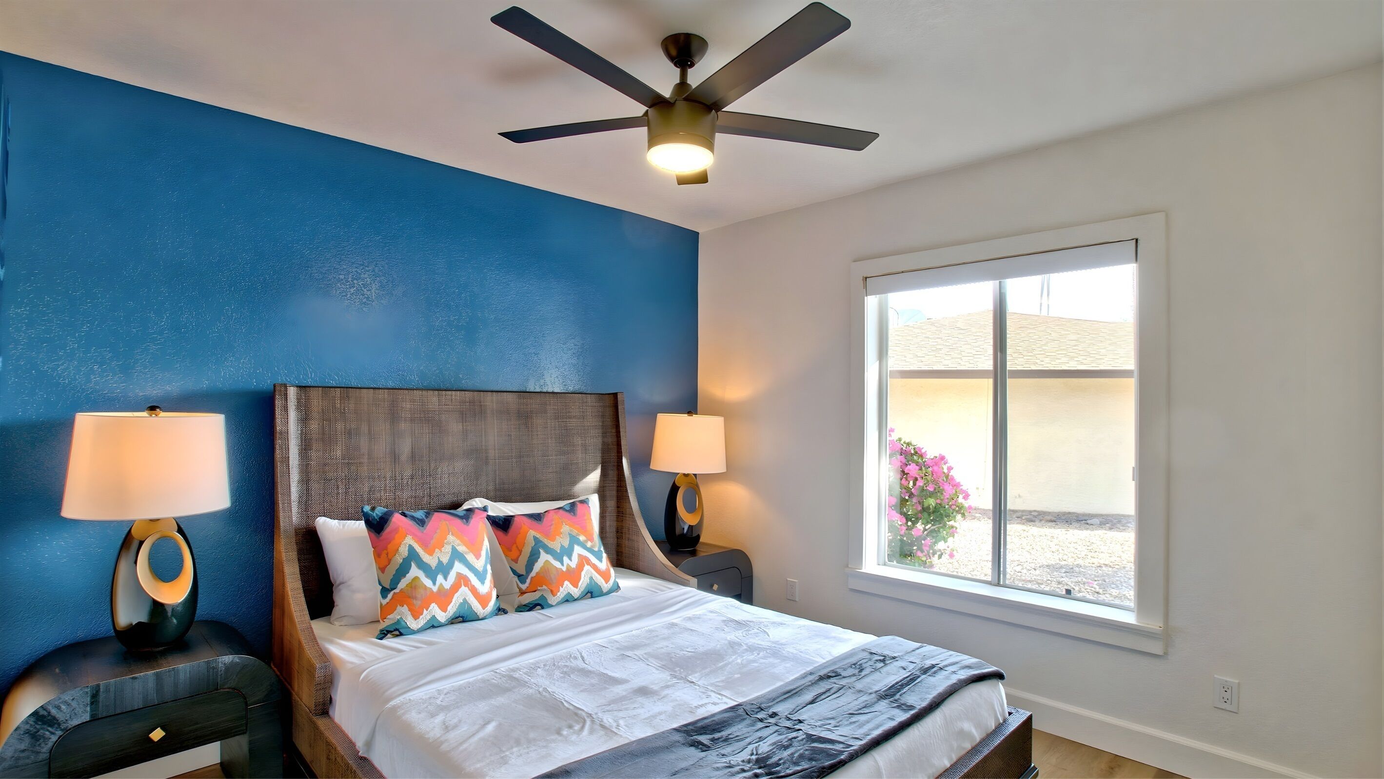 A bedroom with a bed, two bedside tables with lamps, a ceiling fan, and a window with a view.