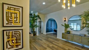 Un interior moderno con suelo de madera, una obra de arte abstracta enmarcada, una consola con espejo y plantas en macetas.
