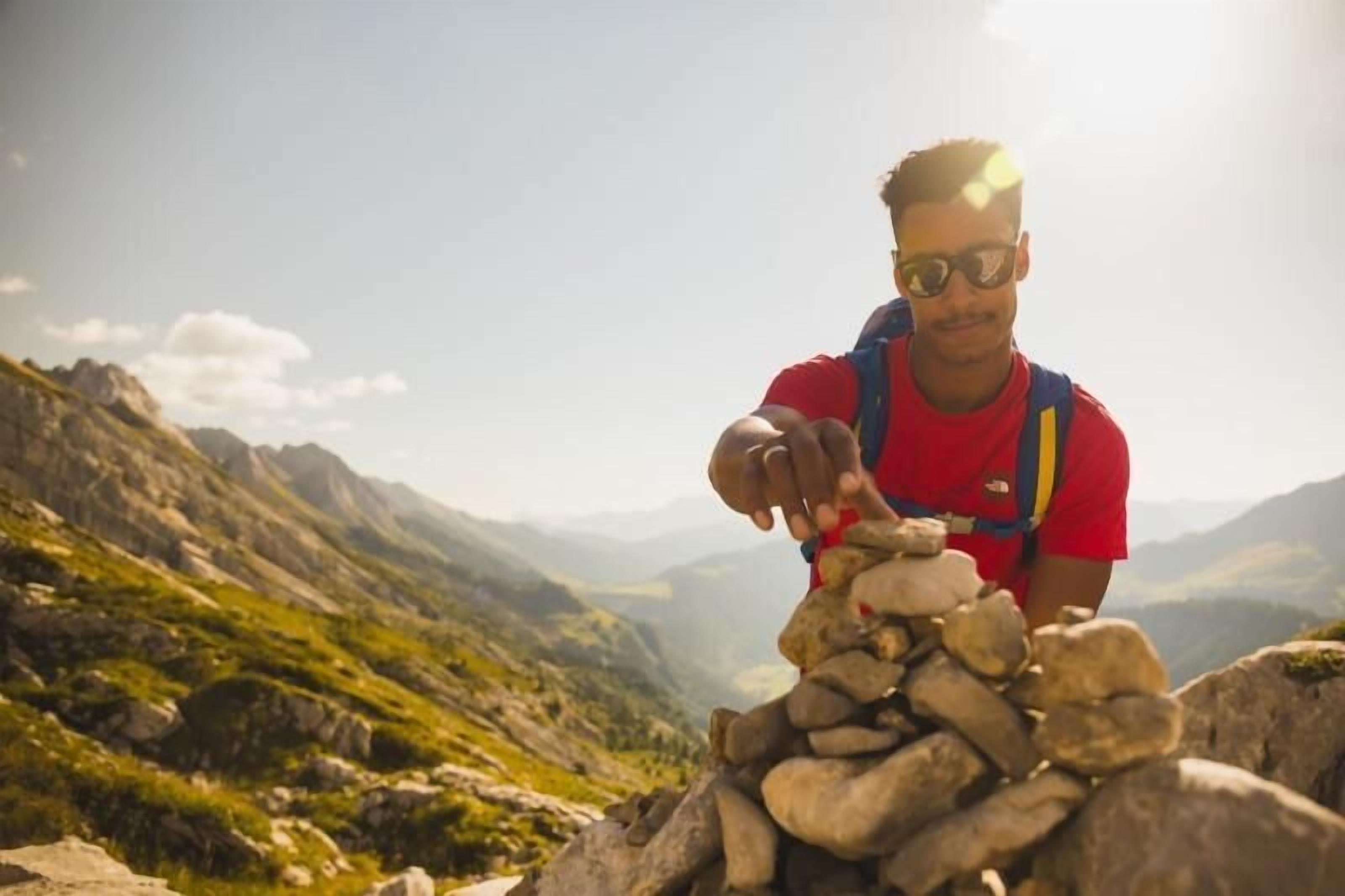 Una persona con camisa roja y mochila azul de pie sobre un saliente rocoso, con un paisaje montañoso al fondo.