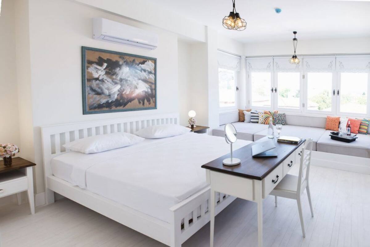 A bedroom with a white bed, a desk, and a large window with a view.