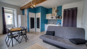 En kompakt opholdsstue med sofa, spisebord, stole, et tekøkken og et vindue med gardiner.