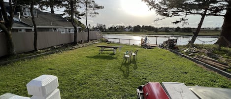 Un patio trasero con césped, una mesa de picnic, una barbacoa roja y vistas a un lago.