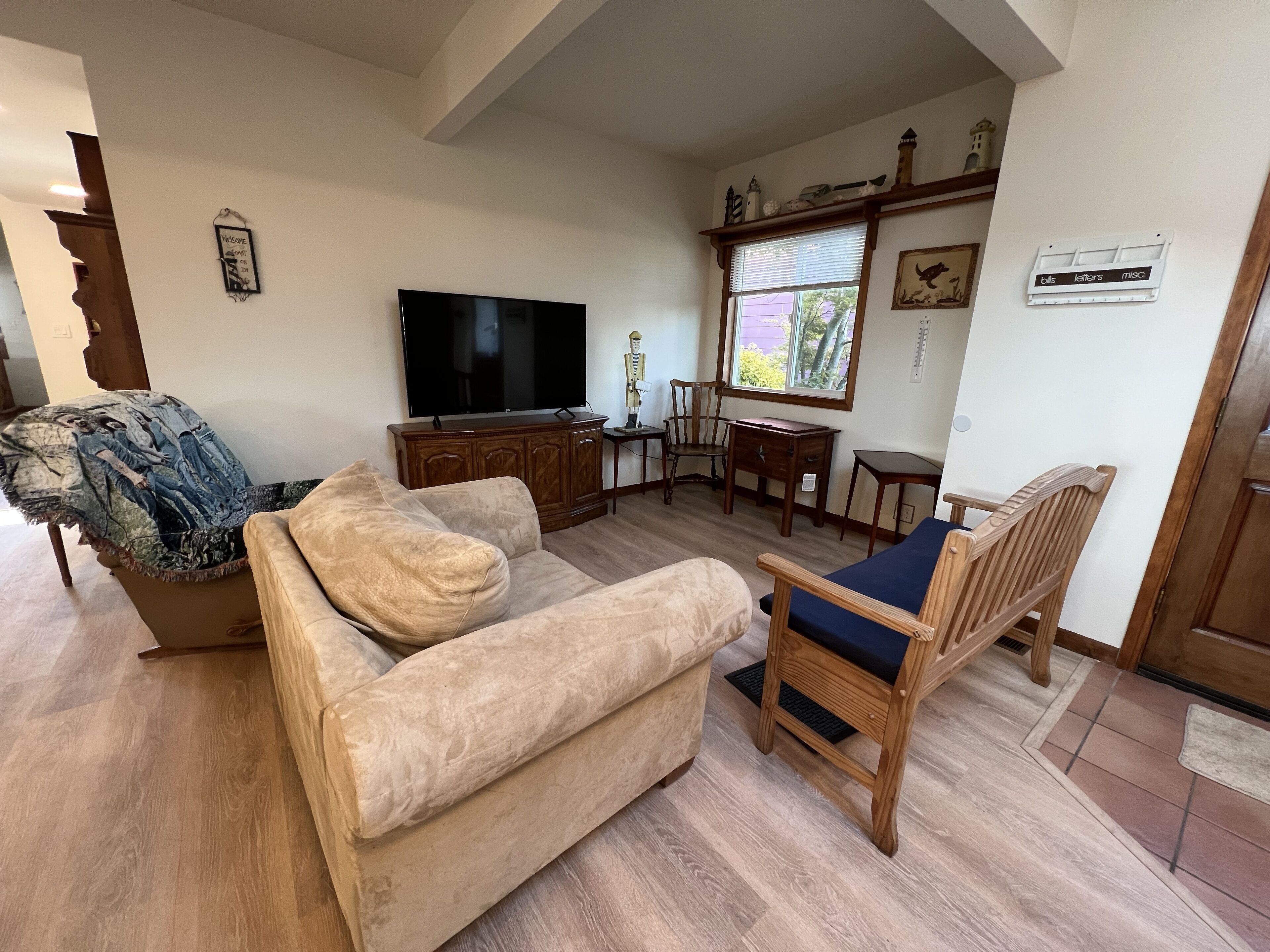 Ein Wohnzimmer mit einem beigen Sofa, einem Holzstuhl mit blauer Kissenauflage, einem Fernseher, einem Holzschrank und einem Fenster mit Jalousien.